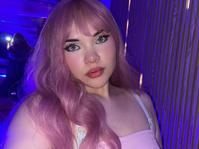 AlayaWillow has pink hair, blue eyes, and full lips with pink lipstick. She's wearing a pink top with white straps, looking directly at the camera with a slight smile.