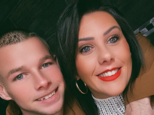 Osmoze has red lipstick and black hair, smiling with bright eyes. Osmoze's white shirt and brown coat are visible. The guy next to Osmoze has short hair and smiles.