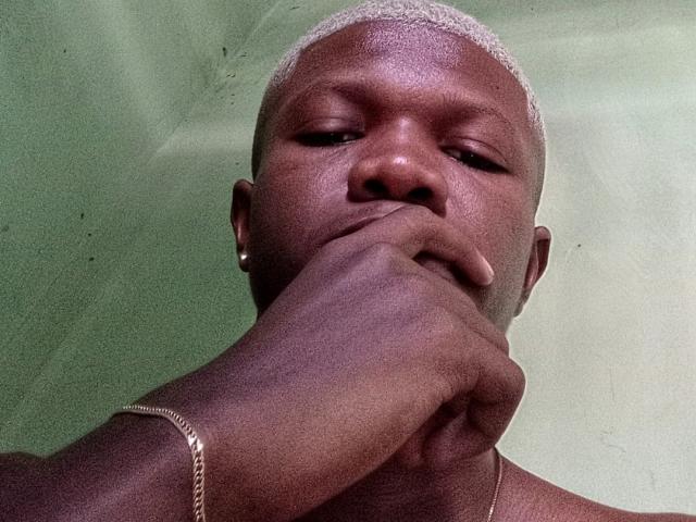 Negobelo's dark brown skin, short white hair, and nose ring, with his hand covering his mouth, wearing a silver bracelet against a green background.