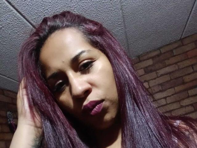 SaraaAdamss with dark purple hair, looking down with a serious expression, slightly parted purple lips, and a hand touching her head, under a textured ceiling.