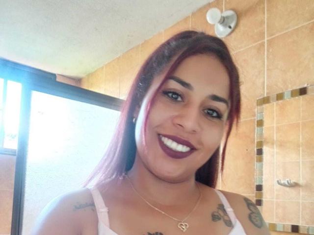 SaraaAdamss hat rote Haare, dunkelvioletten Lippenstift und Tätowierungen an ihren Schultern. Sie trägt ein weißes Tanktop mit einer goldenen Halskette und lächelt in einem Badezimmer..