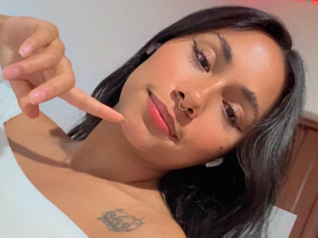 AnaRome has tan skin, black hair, a nose ring, and a tattoo on her left shoulder. She points at her chin with her right index finger.