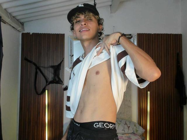 EnergyLove, a young man with curly blonde hair and fair skin, is lifting his white and brown striped shirt to reveal his bare chest. He's wearing a black cap with a white logo, black underwear with "GEORGY" on the waistband, and a black mesh jockstrap hangs on the wooden wall behind him. He's smiling slightly, with a relaxed, confident posture. The lighting is bright, highlighting his toned, slim physique. The image focuses on his upper body and expression.