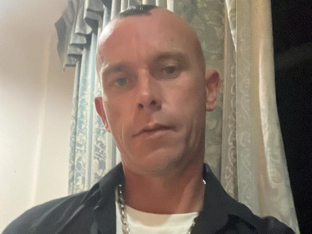 SexyTonyE has a bald head, slight tan, and a chain necklace, wearing a dark shirt over a white tee, with a serious expression, in a slightly blurry photo.