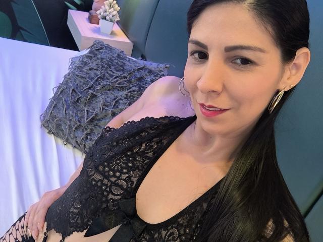 EliseBell, con lunghi capelli neri, indossa una camicetta di pizzo nero che mette in mostra il seno, sorride alla fotocamera, ha le labbra rosa, orecchini a cerchio e siede su un letto con un cuscino grigio..