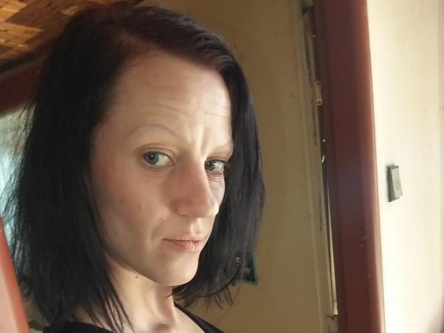 LussyKlara with pale skin, black shoulder-length hair, and blue eyes, showing a slightly confused or serious expression, standing near a wooden door.