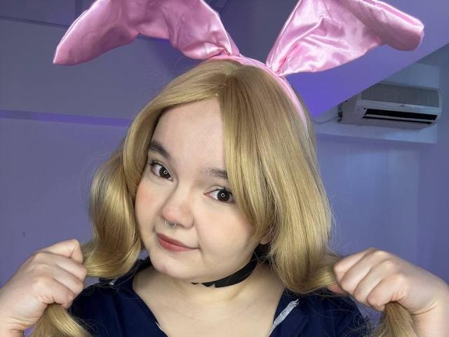 RoseLuv with blonde pigtails, pink bunny ears, and a black choker, holding her pigtails, smiling, showing her nose ring, in a blue top, under purple lighting.
