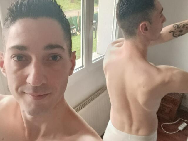 Shirtless FrancaisH with short dark hair, smiling in front, showing back muscles, white towel, tattoo on right arm, standing near window, daylight, visible ear, short brown hair.