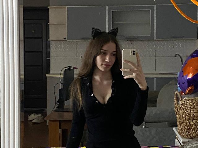 KaiaLein, avec ses longs cheveux bruns, portant des oreilles de chat noires et un haut noir à boutons, décolleté, montrant ses seins dans une photo de profil prise dans un miroir, tenant un iPhone doré..