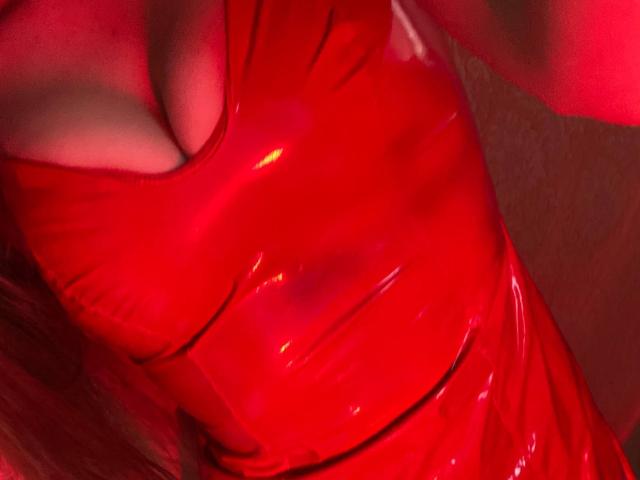 KaiaLein's shiny red latex dress, showing her big, juicy tits with a deep cleavage, reflecting bright red light. Her skin is slightly visible beneath the glossy material.