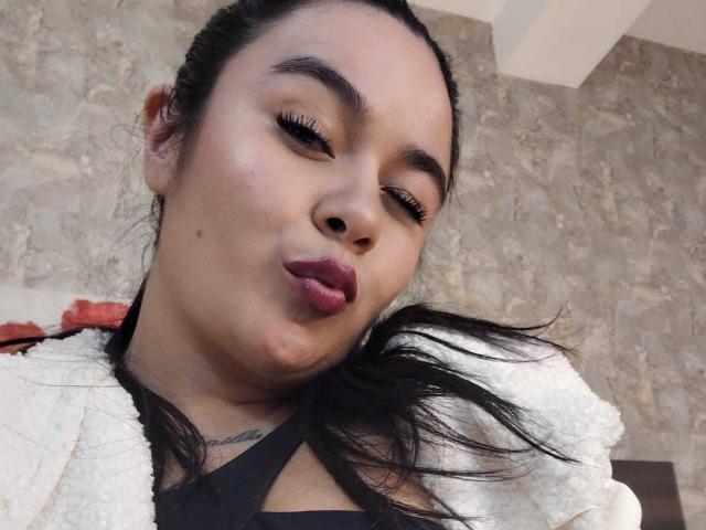 HannahMolly with puffy lips, dark eyelashes, and a small mole on her cheek, wearing a white fluffy jacket, black top, and red backpack, blowing a kiss.