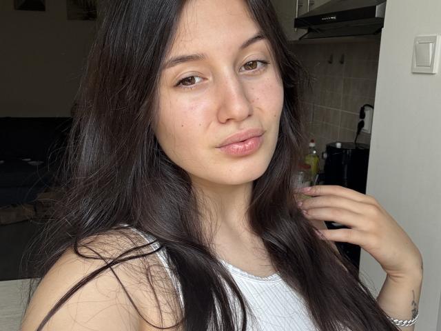LeeaRose with long dark brown hair, brown eyes, pink lips, white tank top, small nose, freckles, hand touching hair, subtle smile, light skin, in a kitchen.