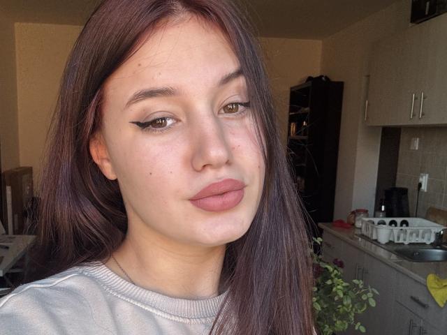 LeeaRose has light brown eyes, black winged eyeliner, pinkish-brown lipstick, and long brown hair. She's wearing a grey sweater, looking slightly to the side with a neutral expression.