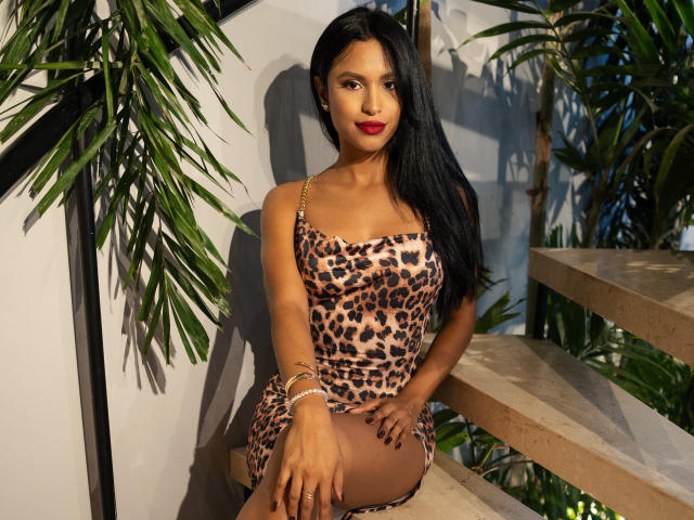 SharonFlorez sits on a concrete staircase, wearing a leopard-print dress with thin gold straps. She has long black hair, red lipstick, and dark eye makeup. Her right hand rests on her thigh, while her left hand is on the stairs. She wears a silver bracelet on her left wrist. The image has strong, natural light, casting shadows on the white wall and green leaves in the background. The composition emphasizes SharonFlorez's confident, relaxed posture and the patterned dress.