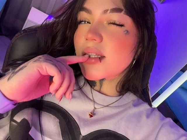 TessaMagic with dark hair, winking, biting finger, nose ring, lip piercing, gray shirt, necklace, tattoos, purple-blue light. Boobless, no visible pussy, no ass. Slightly open mouth.