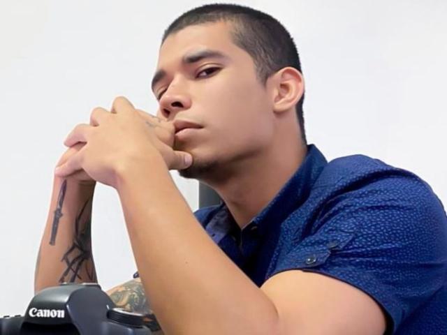 JackBalam, a short-haired dude with tattoos, ponders with his chin on his hands, wearing a blue shirt, camera in front, looking serious and focused.