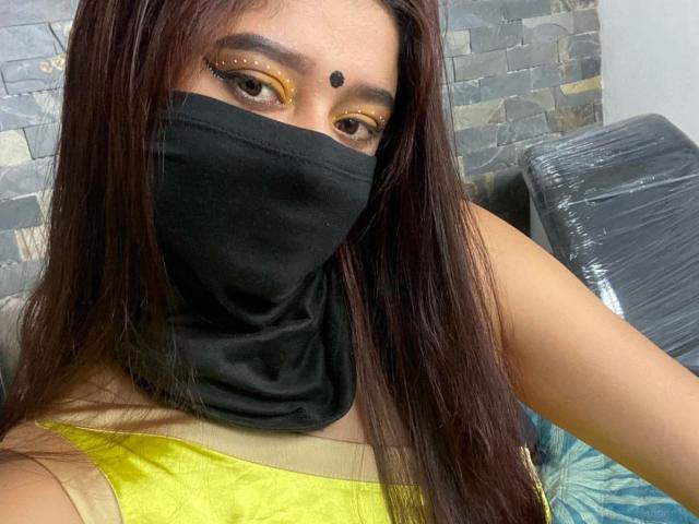 Ananya69 with long brown hair, yellow makeup, black mask, and yellow top, showing her brown eyes and small black dot on her forehead. Her tits are partially visible.