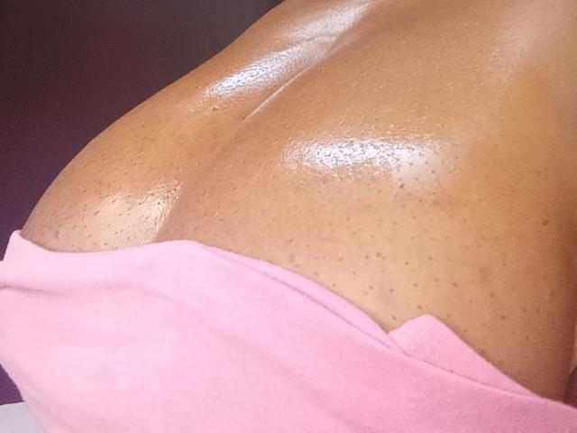 Close-up of Randyxboom's shiny, dark brown, oiled skin on their upper chest and shoulder. The skin has small, visible pores and a slightly textured surface. A pink cloth is partially draped over the lower part of Randyxboom's chest. The light highlights the sheen of the oil, creating reflective, glossy patches. The image focuses on the smooth, oiled skin with a subtle, natural texture. The pink cloth contrasts with the dark skin. The composition emphasizes the texture and shine of the skin.