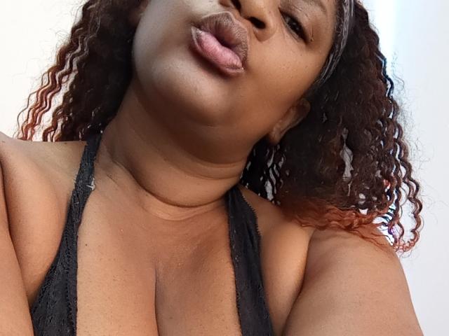 Close-up of Randyxboom with dark brown, curly hair that has red tips. Randyxboom is pouting with slightly parted lips. They have a medium-dark skin tone and are wearing a black lace halter top that reveals cleavage. The texture of Randyxboom's hair is wavy and slightly frizzy. The image focuses on Randyxboom's face and upper chest, with a slight upward angle. The background is blurred and light-colored. The image emphasizes Randyxboom's facial expression and hair texture.