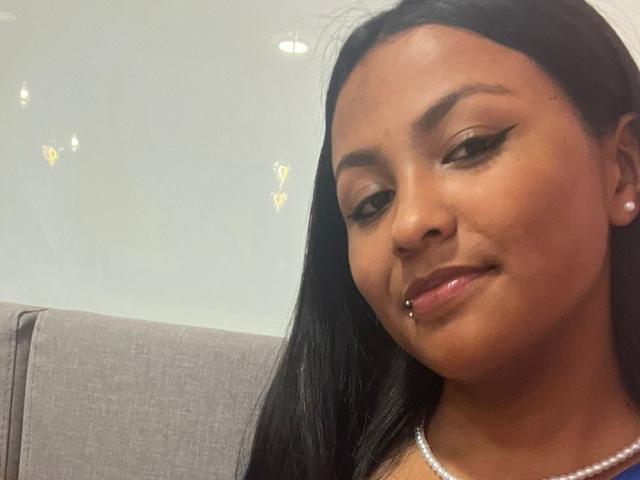 NaomiRios has dark brown skin, black straight hair, and a septum piercing. She's wearing a pearl necklace, has winged eyeliner, and a subtle smile. She's sitting on a grey chair.