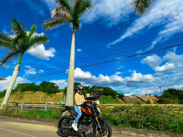 KimmyK, che indossa una giacca gialla e un casco nero, è seduta su una moto arancione, parcheggiata su una strada, con due alti alberi di palma e un cielo azzurro brillante..