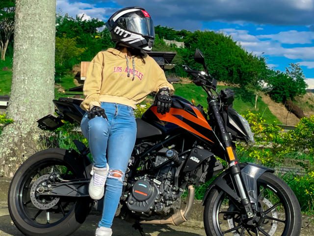 KimmyK appuyée contre une moto noire et orange, portant un casque, un sweat à capuche beige, un jean bleu déchiré, des baskets blanches et des gants noirs, avec un arbre et de la verdure en arrière-plan..