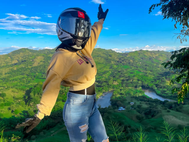 KimmyK with a helmet, yellow hoodie, and ripped jeans, pointing up with her right hand, standing on a green hill, blue sky, and a river in the background.