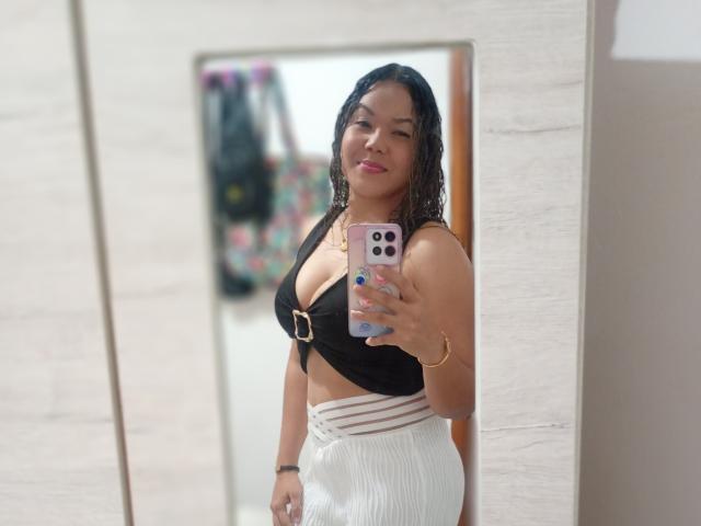 Estrellya with wet black hair, wearing a black crop top with gold buckle and white high-waisted skirt, holding pink phone, showing her tits and a cute smile in mirror.