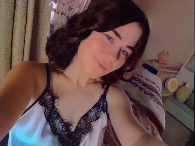 ToryTayler with wavy brown hair, fair skin, blue eyes, and a small smile, wearing a lacy black and white camisole, showing her tits in a selfie.