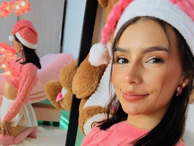 KimmyK, cute Latina woman with dark hair, wearing pink Santa hat, pink sweater, white skirt, showing her small pink-ass in mirror selfie, smiling, pink earrings, reflecting on wall.
