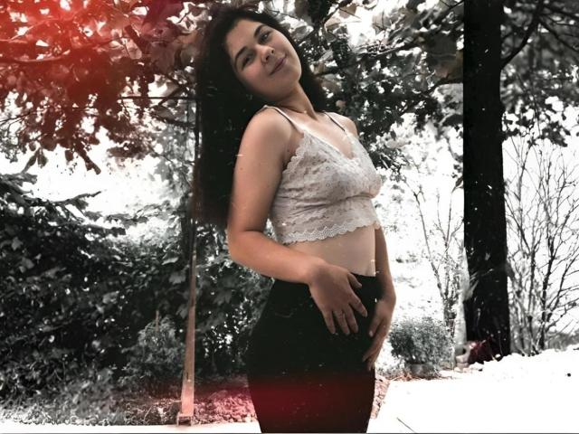 ToryTayler stands in a snowy forest, wearing a white lace crop top and black high-waisted pants, with long dark hair and a slight smile, showing her tits.