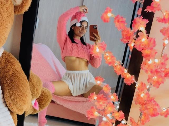 KimmyK, portant un chapeau rose, un crop top et une jupe blanche, assise les jambes écartées, montrant sa vulve, dans un selfie devant un miroir avec des fleurs roses et des lumières, et un ours en peluche marron..