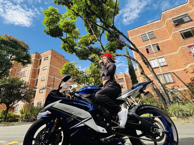 KimmyK sits on a blue and white motorcycle, wearing black pants, a black crop top, and a red cap. She's holding a phone in her right hand, with a tree and red-brick apartment building in the background. The motorcycle has a sleek, modern design with angular lines and a shiny finish. Bright sunlight casts shadows, highlighting the motorcycle's details and KimmyK's casual, relaxed posture. The image is sharp, with vibrant colors and a clear, sunny sky.