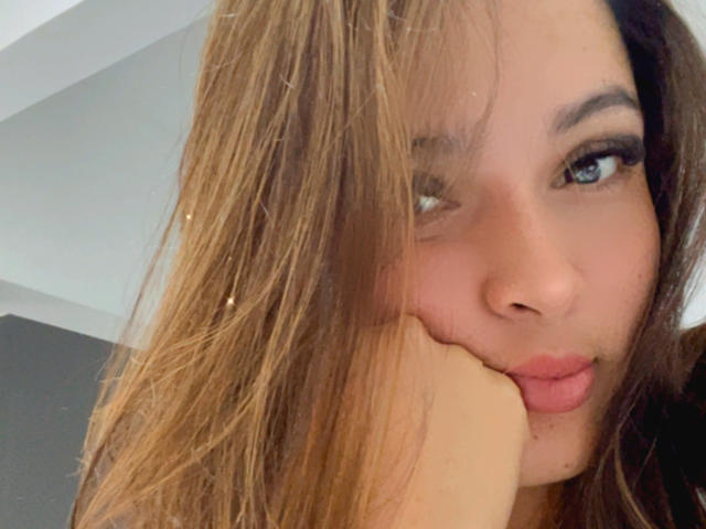 Gabrielafoxxy has pouty lips, a cute nose, and is biting her index finger with her blue eyes looking sideways. Her brown hair is slightly messy.