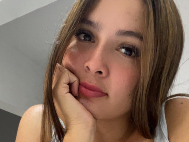 Gabrielafoxxy with brown hair, blue eyes, and pink lips, resting her chin on her hand, looking up with a slight smile, showing her nose and small, freckled cheeks.