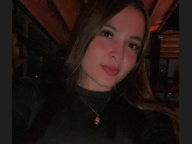Gabrielafoxxy with long brown hair, wearing a black turtleneck, subtle smile, and a gold necklace with a red pendant, in dim, warm lighting.