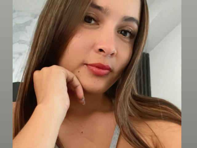 Gabrielafoxxy with long brown hair, light skin, small moles on her chin, pink lipstick, and a slight smile, resting her cheek on her hand. Boobs not visible.