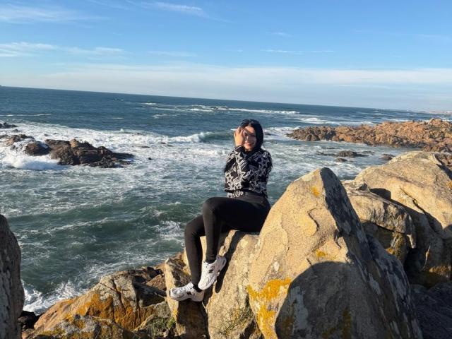 IsabellaJames, with black hair and black patterned shirt, sits on a rock by the ocean, wearing black pants and white sneakers, with a blue sky and waves behind.