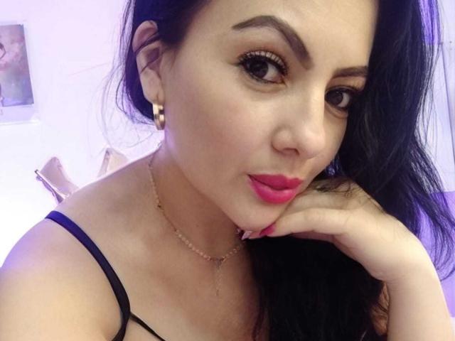 JakyKidman with dark hair, pink lipstick, black strappy top, gold earrings, and necklace, resting chin on hand, looking at camera.