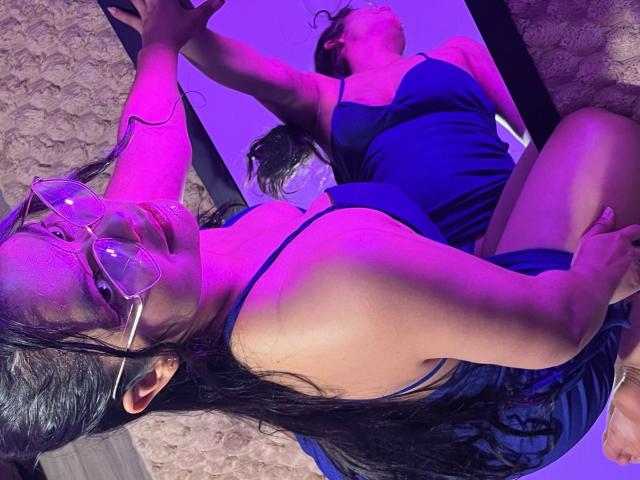 AmaiaSaenz with glasses, blue dress, and long black hair, kneeling, arms raised, purple lighting, reflected in a mirror, showing her tits and bare ass, on a textured surface.