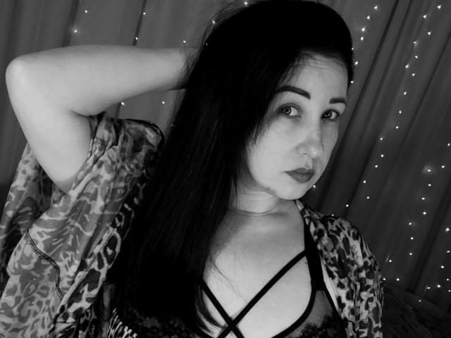 JillSilvies, con lunghi capelli scuri, un accappatoio a stampa leopardo, lingerie nera con spalline sottili, una mano tra i capelli, che guarda la fotocamera, rossetto nero e luci a stringa sullo sfondo..