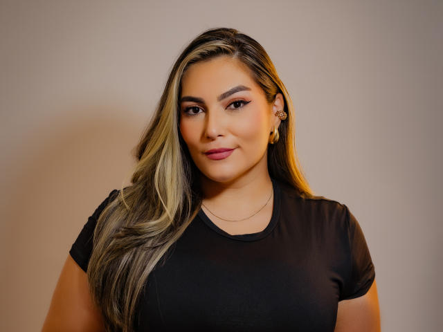 LadyVesper with long blonde and dark brown hair, wearing a black shirt, smiling, has big tits, gold earrings, and a small necklace, against a plain background.