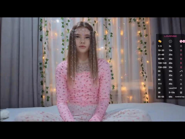 KirstieDevost with long braided hair, wearing a pink floral long-sleeve top and white pants, sitting on a bed with fairy lights in the background. Her tits are small.