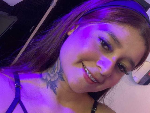Ananya69 with purple-lit face, brown hair, black headband, smiling, showing metal teeth, and floral tattoo on her neck, wearing a black strappy top.