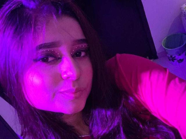 Ananya69 has pink-lit brown hair, purple eyeliner, and a pouty expression. She's wearing a red shirt, with purple and pink lighting on her face. Her eyes are half-open.