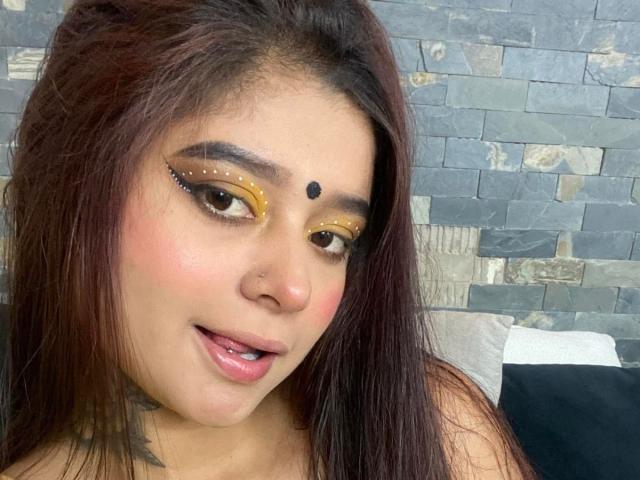 Ananya69 has brown hair, brown eyes, yellow eyeliner, a black dot on her forehead, and a nose piercing. She's sticking her tongue out slightly. Her tits are not visible.