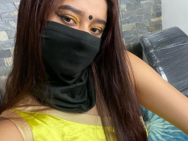 Ananya69 with long brown hair, black mask covering nose and mouth, yellow top, dark eye makeup, and a black dot on her forehead. Her eyes are looking at the camera.