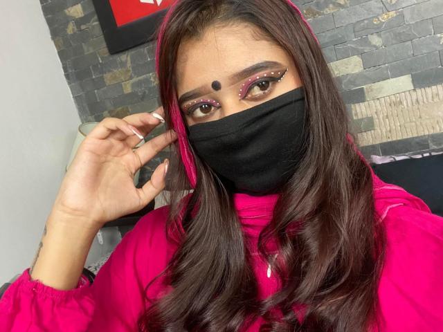 Ananya69 with long brown hair, pink eyes, black mask, red headscarf, pink eyeliner, and pink top, touching her face. Boobs not visible, background ignored.