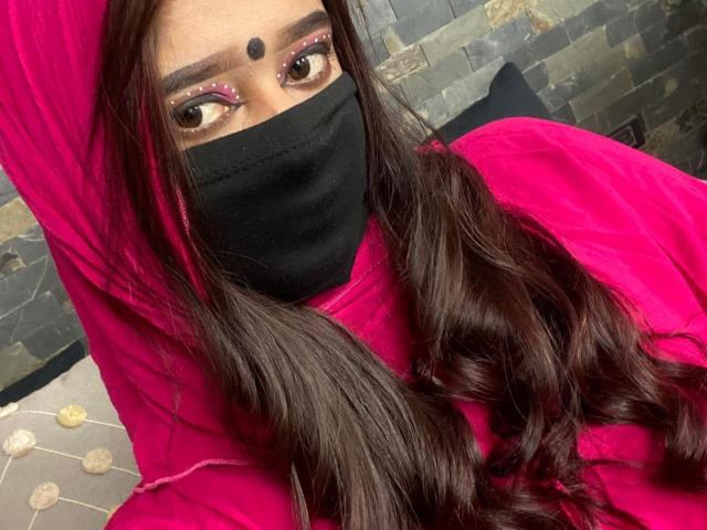 Ananya69 with long dark hair, pink eyes, black mask, and pink outfit, raising arm, showing black dot on forehead, pink eyeshadow, and pink eyebrows. Boobs partially visible.