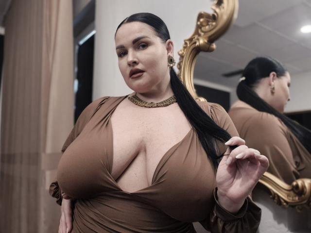 GeorgiaLerox with large tits, brown dress, deep cleavage, black ponytail, gold necklace, looking down, holding hair, reflective gold mirror, soft lighting, confident expression.